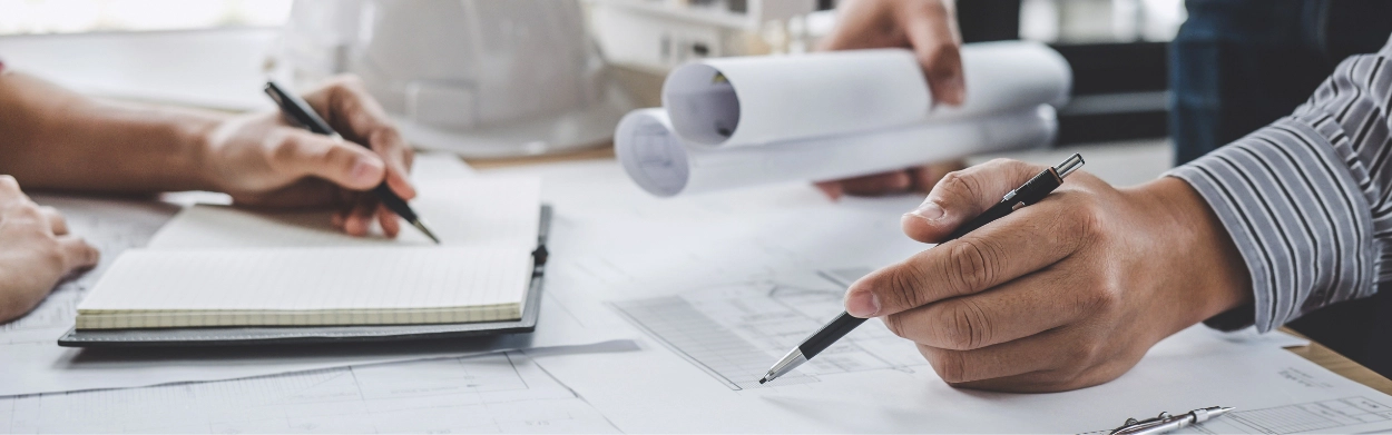 Two people working at a desk with architectural plans for Pre-Engineered Metal Buildings, one holding rolled blueprints and the other writing notes, with papers and a hard hat visible.