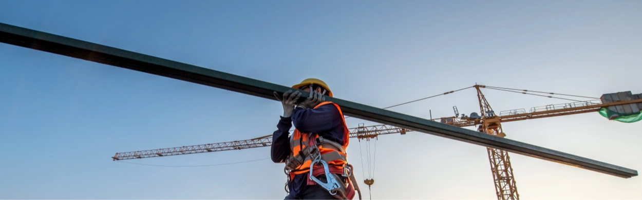 Construction worker in safety gear carries a metal beam on a building site with cranes in the background, showcasing the process of assembling Pre-Engineered Metal Buildings against a clear sky.