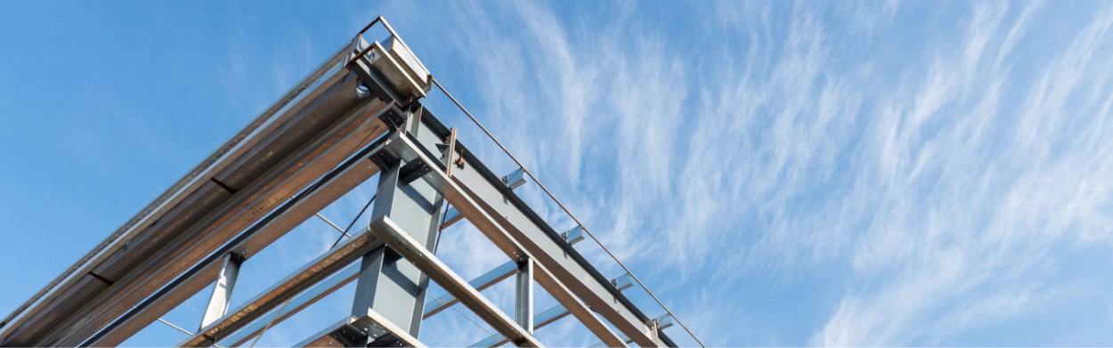 Steel framework of a Pre-Engineered Metal Building under construction rises against a blue sky with scattered clouds.