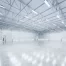 Large empty warehouse with high ceilings and metal beams typical of commercial metal buildings, bright lighting, and polished concrete floors. Walls are white with several doors and yellow-black striped pillars.