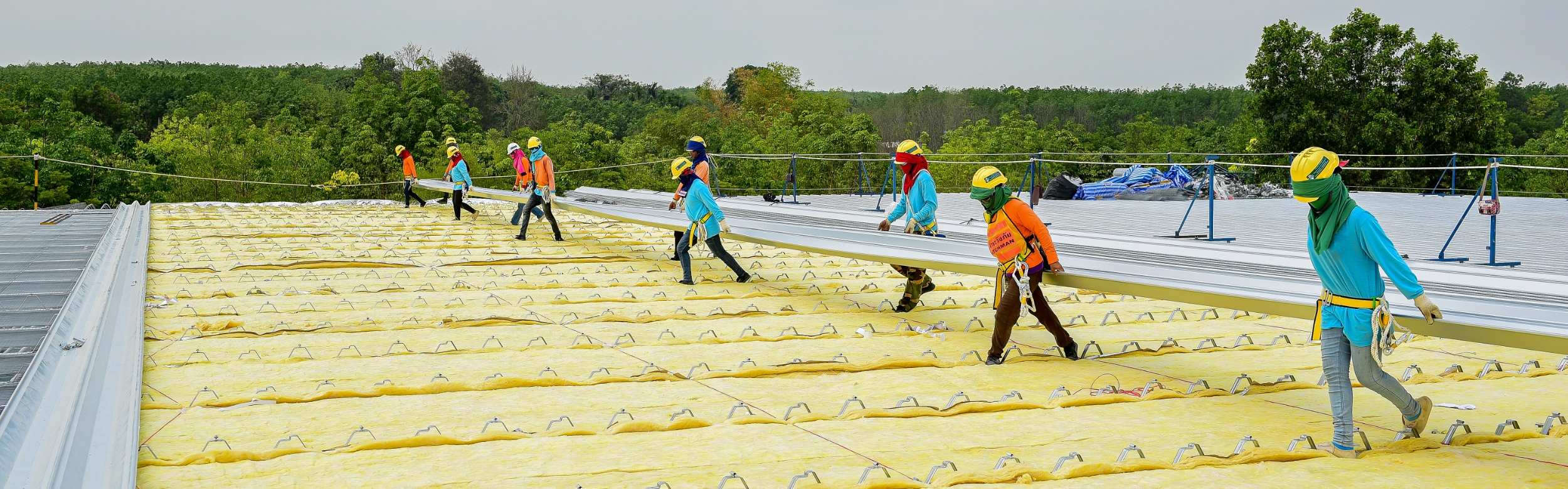 Workers in protective clothing install insulation panels and metal sheets as part of a Metal Building Re-Roof on a large rooftop, surrounded by trees and greenery under an overcast sky.