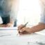 Two people review architectural blueprints on a table, focusing on manufacturing construction details; one writes with a pen while the other stands in the background near a safety helmet, emphasizing risk management.