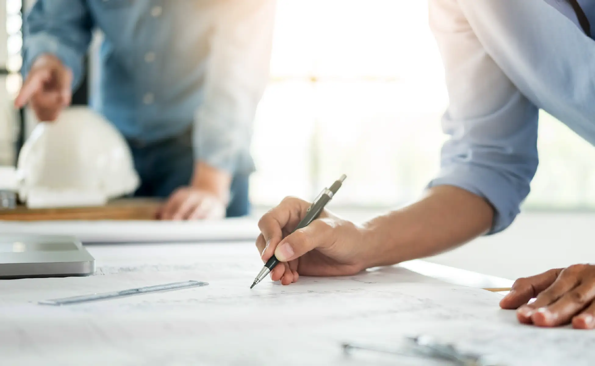 Two people review architectural blueprints on a table, focusing on manufacturing construction details; one writes with a pen while the other stands in the background near a safety helmet, emphasizing risk management.